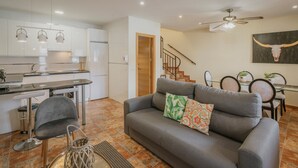 A modern kitchen with white cabinets, a dining area with a glass table, and a gray sofa with patterned cushions.