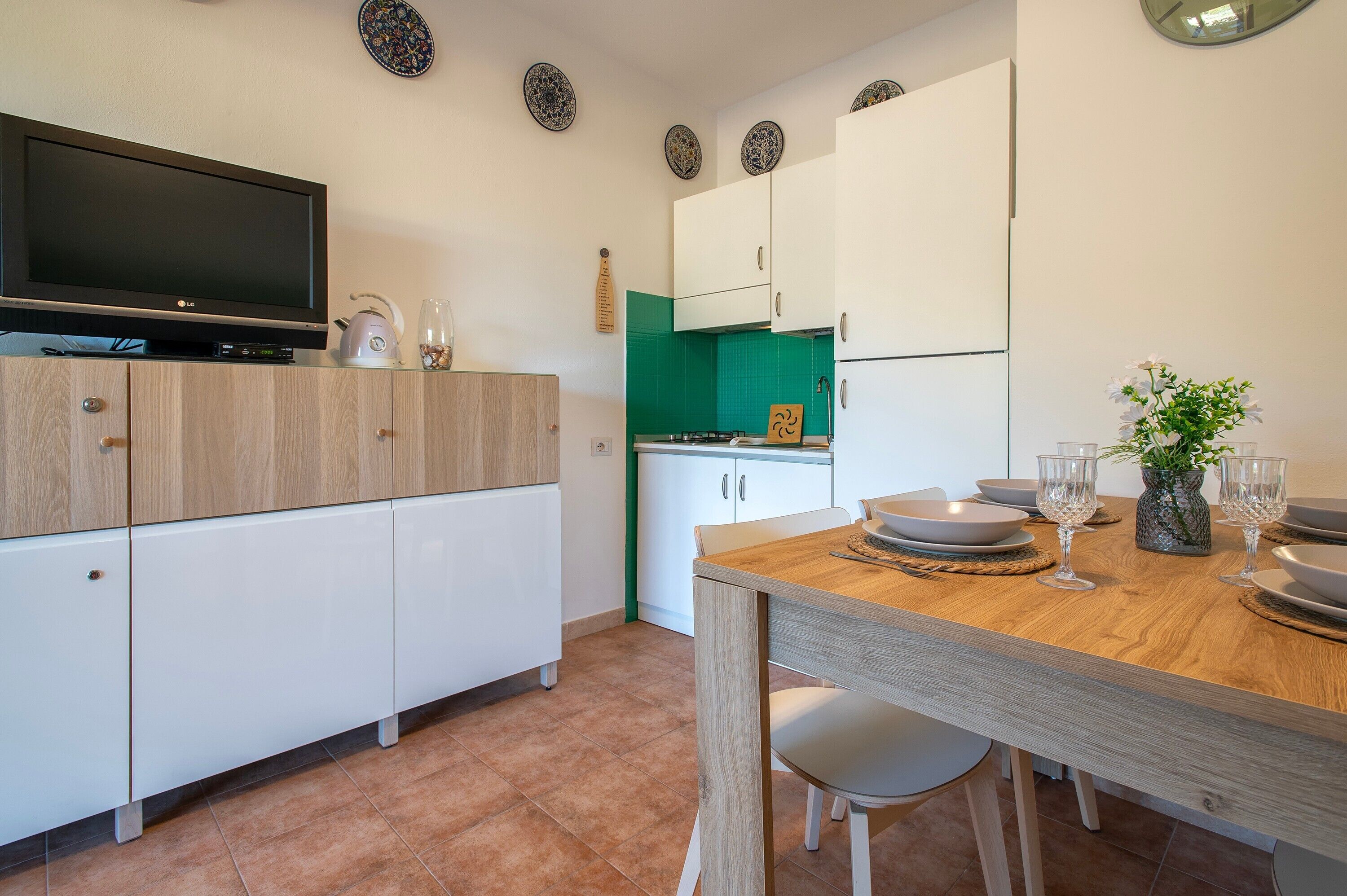 Una cocina moderna con televisor de pantalla plana, armarios blancos, una mesa de comedor con sillas y un salpicadero verde.