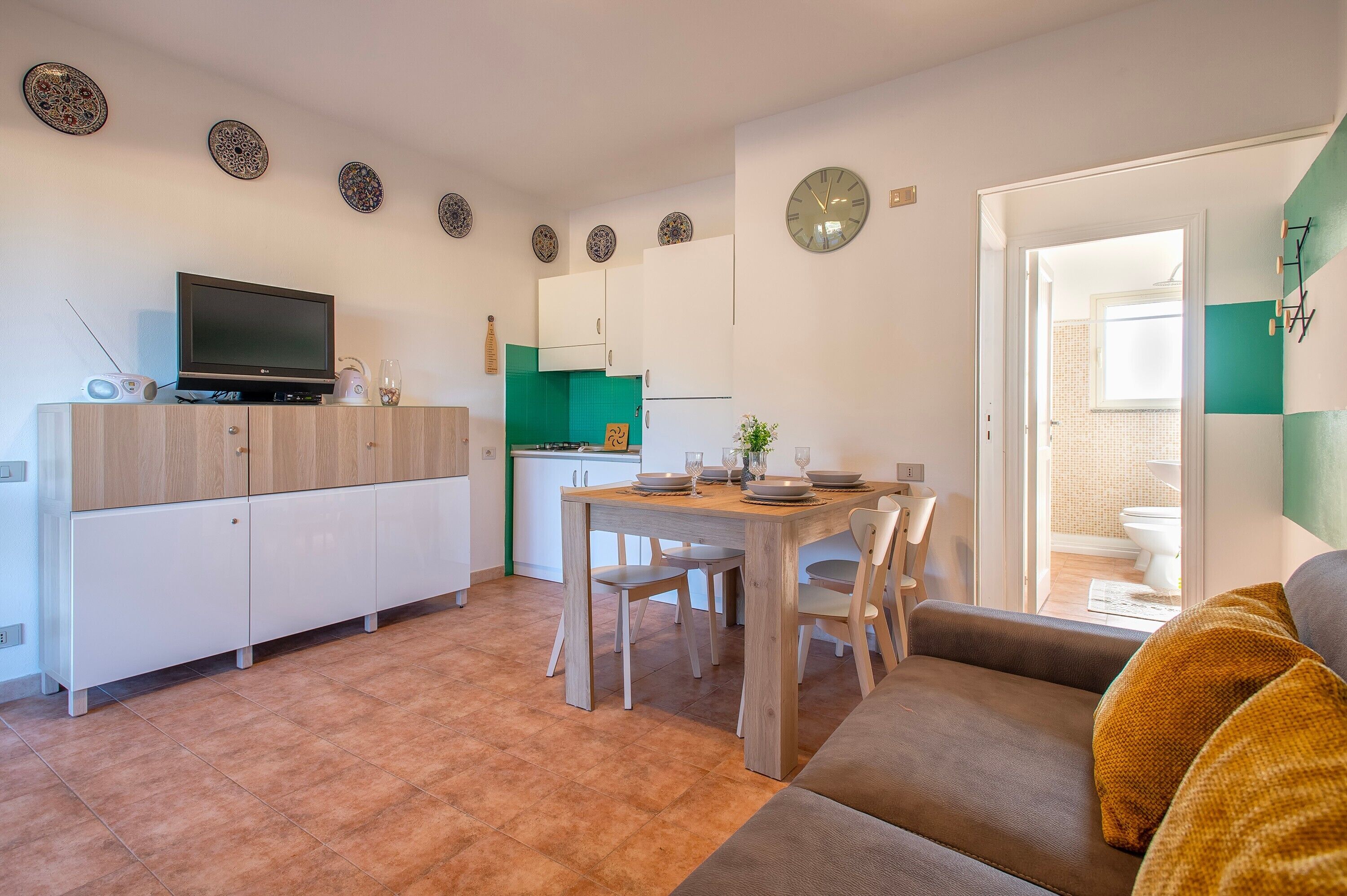 Une cuisine moderne avec des meubles blancs, une table à manger avec des chaises, un téléviseur posé sur un meuble, et une vue sur la salle de bain.
