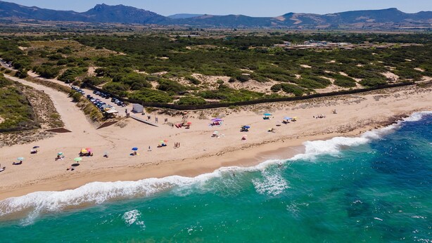 Vista aérea de una playa con una fila de coches aparcados, sombrillas y gente.