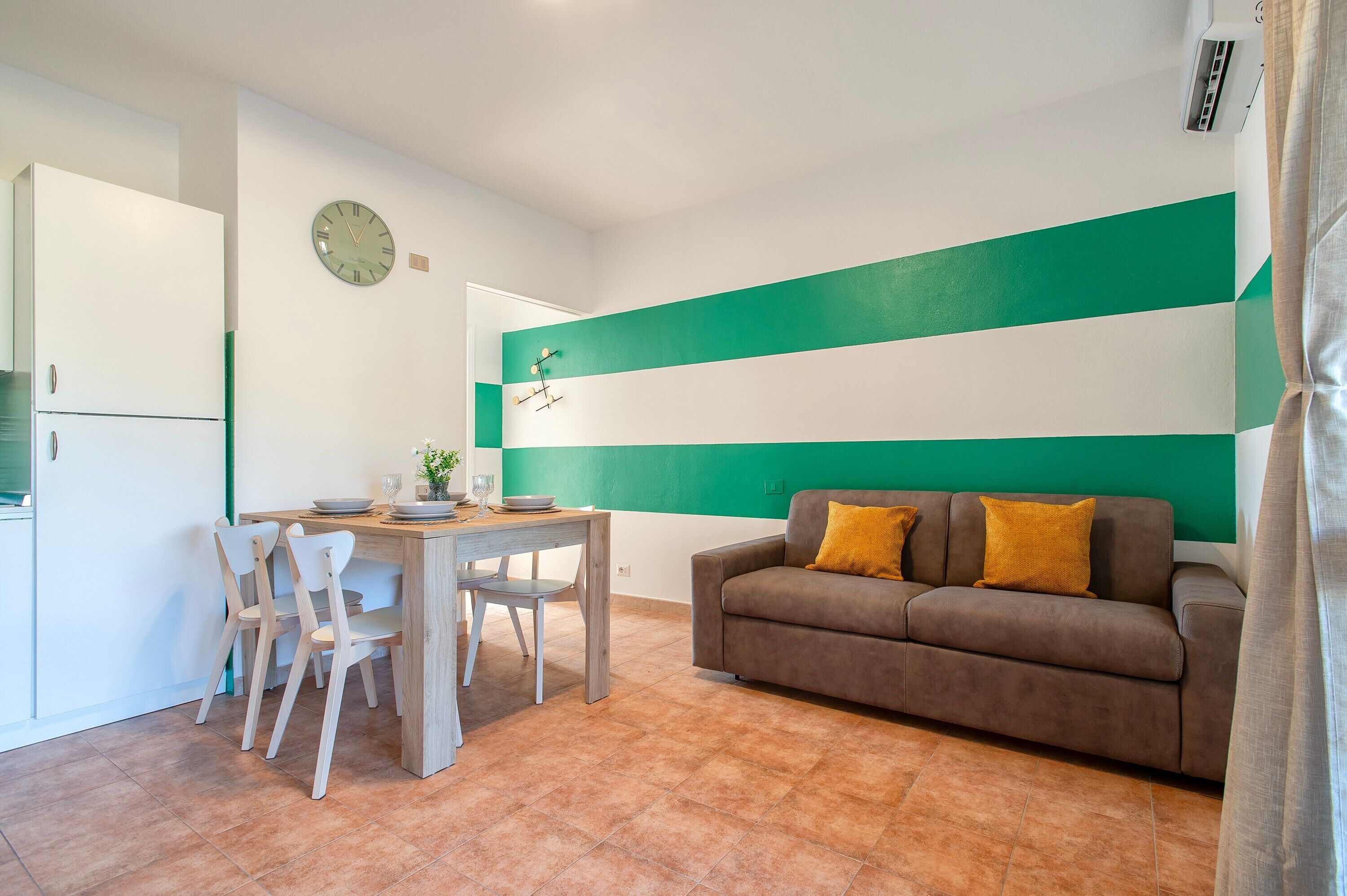 Ein Wohnzimmer mit einer Couch, einem Esstisch mit Stühlen, einer Uhr an der Wand und einem grün-weißen Streifenmuster an der Wand.