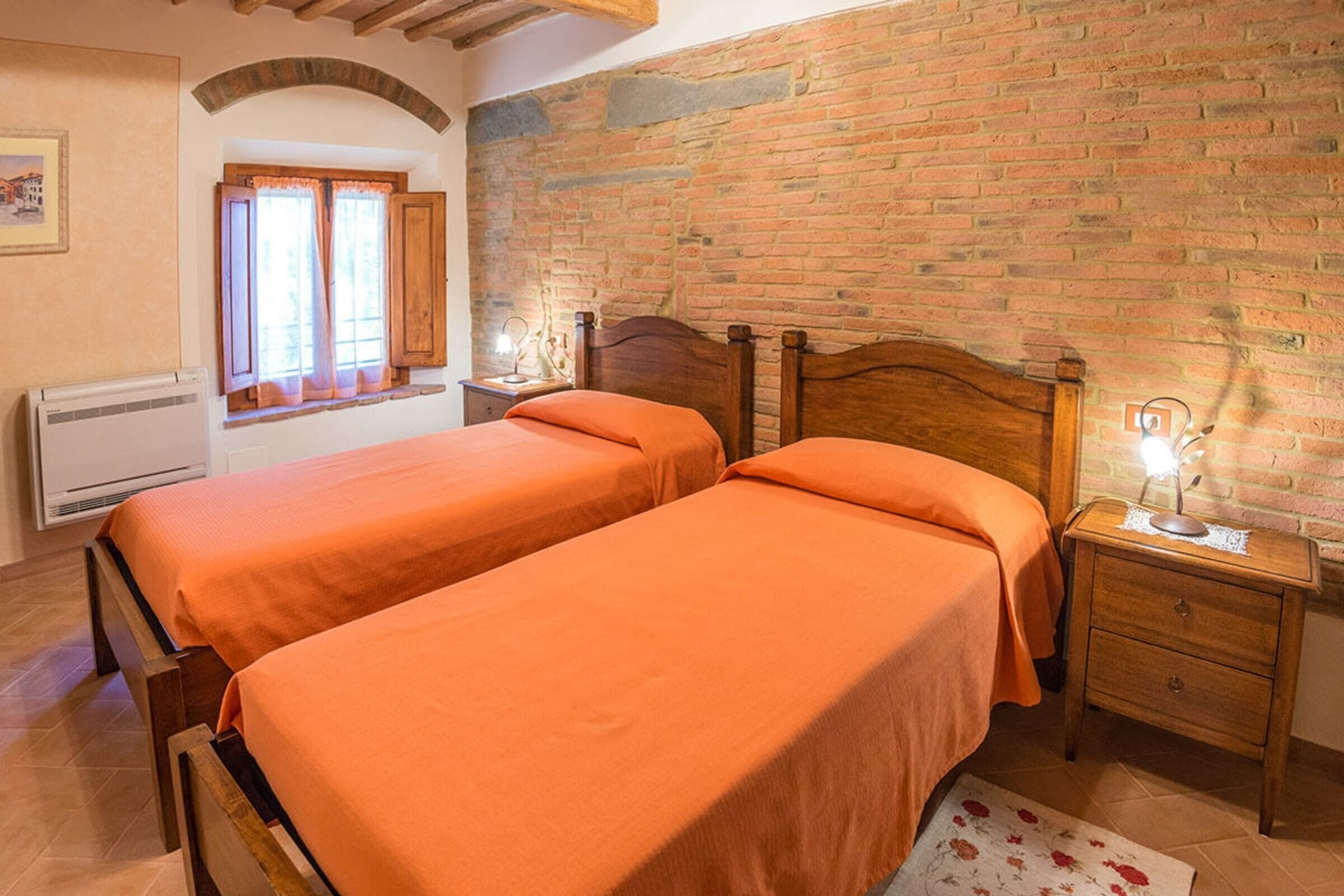 A bedroom with two beds, a brick wall, a window with curtains, a bedside table with a lamp, and a radiator.