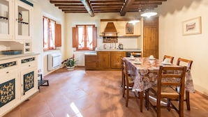 Zona de menjador amb una taula preparada per a quatre persones, caires de fusta, cuina amb fogó i armaris, i finestra amb cortines.