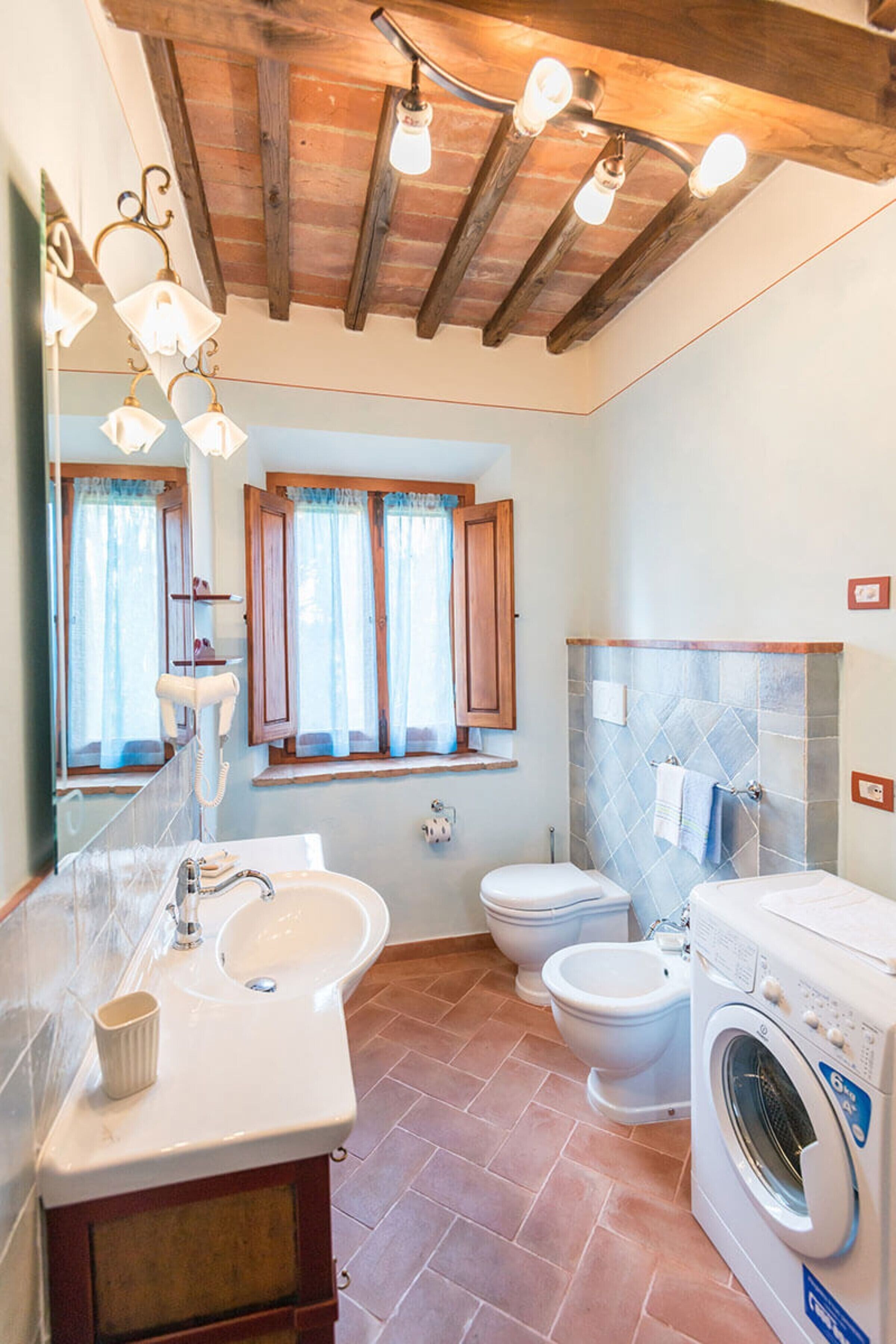 Une salle de bain équipée d’un lavabo, d’un miroir, de deux toilettes, d’un lave-linge et d’une fenêtre avec des rideaux.