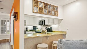 A modern kitchen with white cabinets, a central island with stools, a microwave, and a decorative panel above the island. - Steps Away from the Pool, Beach, Restaurants, Village, and Pier! (Saint Simons Island)
