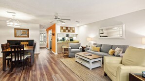 A modern living room with a dining area, kitchen, and a ceiling fan. - Steps Away from the Pool, Beach, Restaurants, Village, and Pier! (Saint Simons Island)
