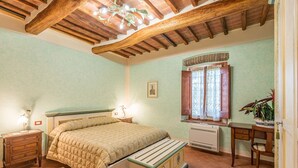 Une chambre à coucher comprenant un lit, une tête de lit en bois, des tables de chevet, un radiateur, une fenêtre avec des rideaux, un bureau et une chaise.