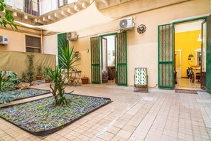 Un patio con suelo de baldosas, plantas en macetas, una pequeña zona de jardín y un comedor con sillas y una mesa.
