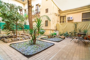 Un patio con suelo de baldosas, plantas en macetas, un árbol y una mesa con sillas.