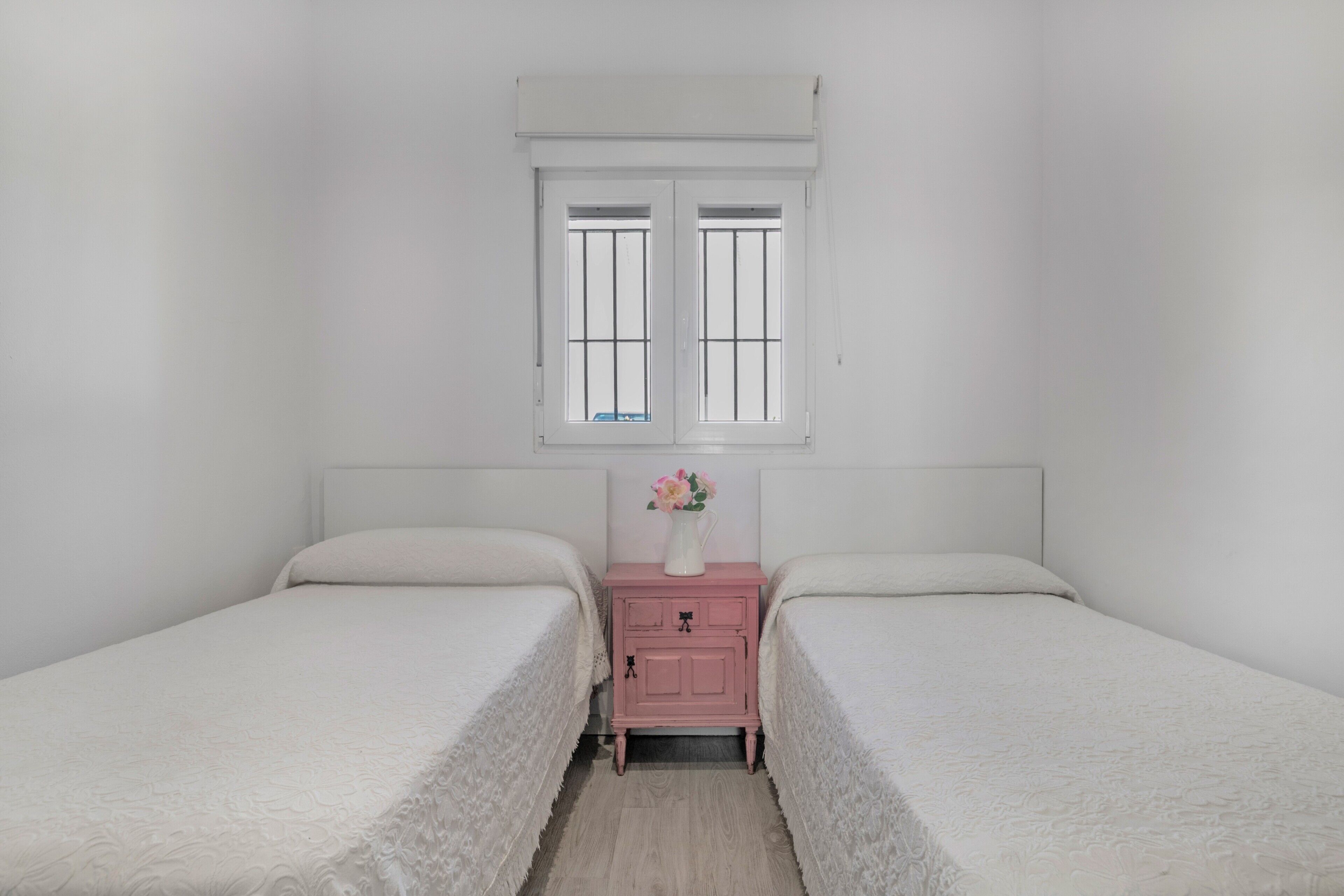 A bedroom with two beds, a nightstand with a vase, and a window with bars.