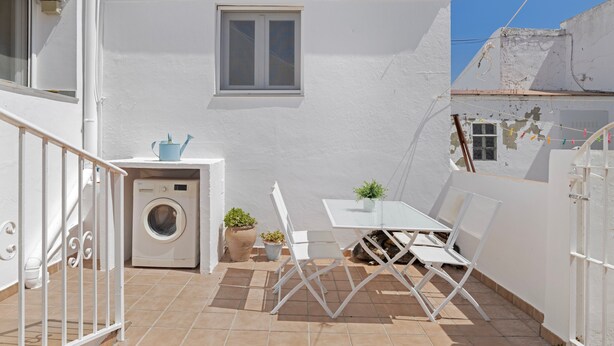 A small, white-walled courtyard with a washing machine, two potted plants, a table with chairs, and a window.
