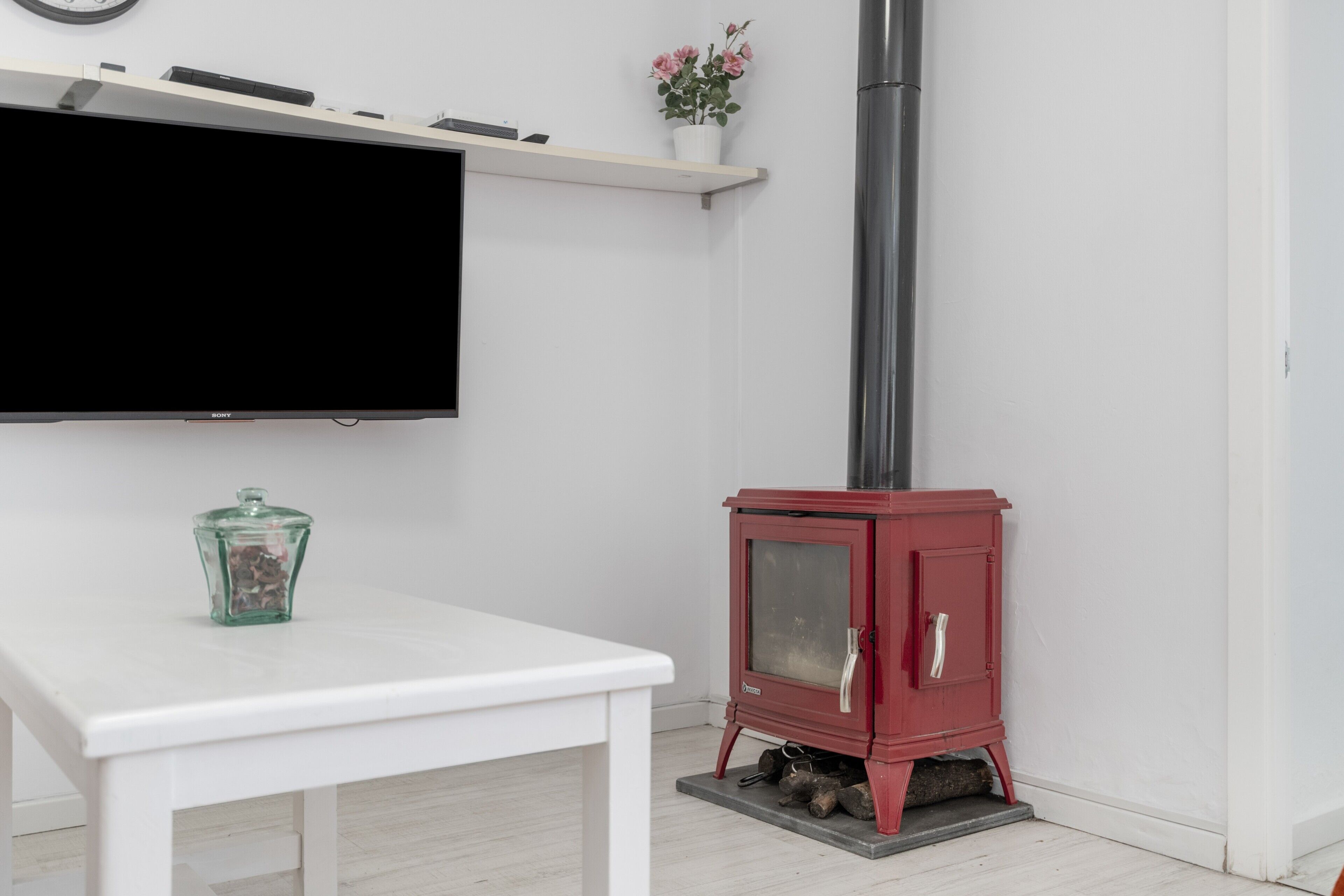 A room with a white table, a red wood-burning stove, a mounted TV, and a small potted plant.