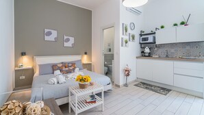 A modern hotel room with a bed, bedside tables, a small white table with a basket of flowers, a kitchen area with a microwave, and a bathroom visible through an open door. - Apartment 'I Sogni Di Ludo' with Shared Terrace, Wi-Fi and Air Conditioning (Siracusa)