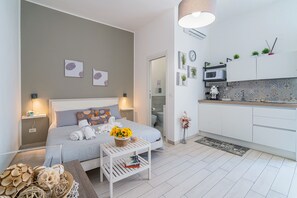 Une chambre d’hôtel moderne, équipée d’un lit, de tables de chevet, d’une petite table blanche agrémentée d’un panier de fleurs, d’un coin cuisine avec un four à micro-ondes et d’une salle de bain visible à travers une porte ouverte.