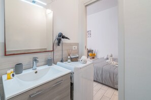 A modern bathroom with a large mirror, a sink, a hairdryer, and a bed with a gray blanket.