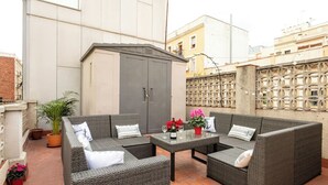 An outdoor seating area with wicker furniture and a small table, surrounded by potted plants and a decorative fence.