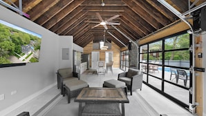 A spacious living area with a high ceiling, a ceiling fan, and large windows offering views of the outdoors.