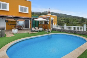 A swimming pool with a patio area, a small building with a tiled roof, and a view of a hilly landscape.