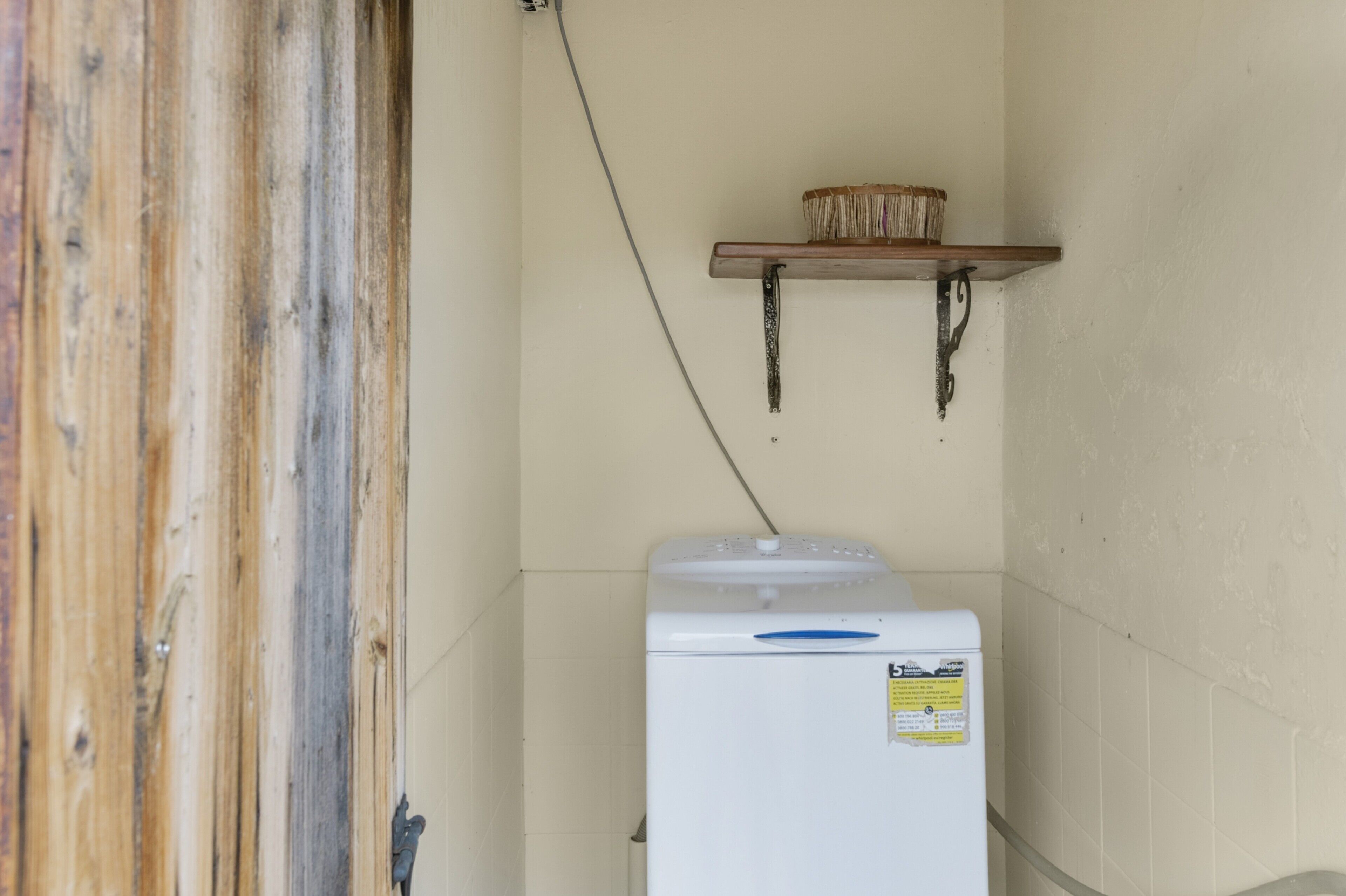 A compact room with a white refrigerator, a wooden shelf with a basket, and a wall-mounted electrical outlet.