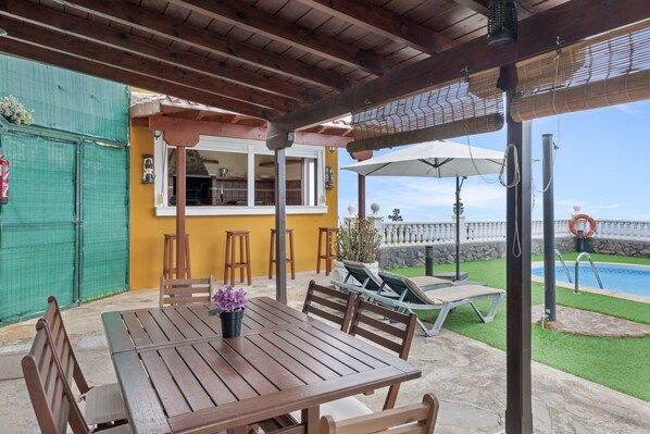 A covered patio with a wooden table and chairs, a bar area, and a view of a swimming pool.