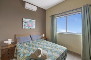 A bedroom with a bed, a nightstand, a wall-mounted air conditioner, a window with curtains, and a painting on the wall.