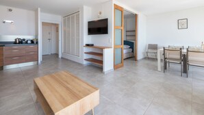 A modern, well-lit room with a wooden table, a TV mounted on the wall, a bed with a blue headboard, and a dining area with chairs. - 2-Br Seafront Apartment in Cannes (Cannes)