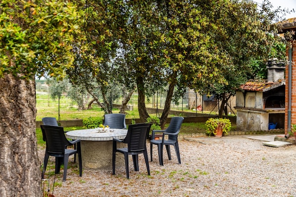An outdoor patio with a round table and four chairs, surrounded by trees and a brick oven.