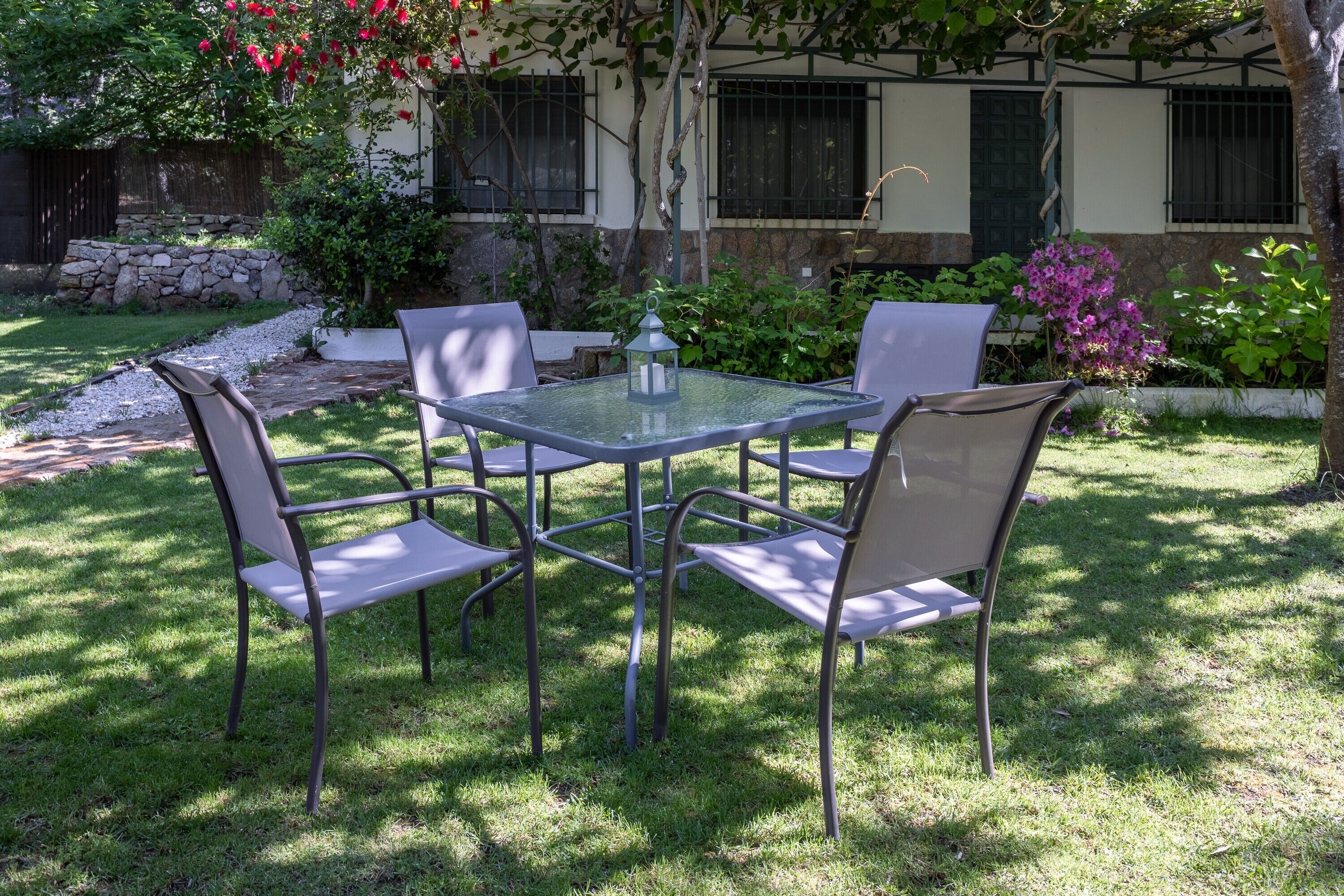 A patio with a glass table and four chairs, surrounded by a garden with various plants and a stone wall.