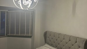 Een slaapkamer met een gestoffeerd hoofdeinde, een bed met een patroon, een ronde lamp en een raam met jaloezieën.