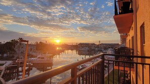A marina with boats docked, a sunset in the background, and a building with a balcony overlooking the water.