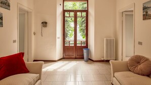 Ein Zimmer mit einer großen Holztür, zwei beigen Sofas, einem roten Kissen, einem Heizkörper und einem Fenster mit Blick auf Bäume.
