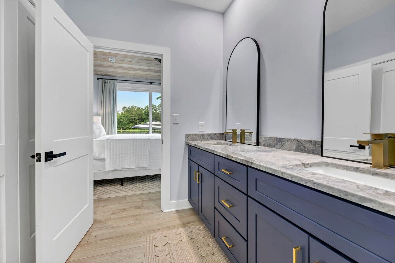 A modern bathroom with a marble countertop, a large mirror, and a view of a bedroom with a bed and window.