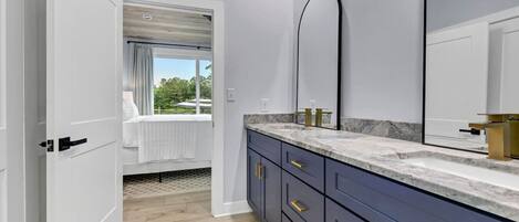 A modern bathroom with a marble countertop, a large mirror, and a view of a bedroom with a bed and window.