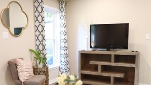 A living room with a glass coffee table, a TV on a wooden stand, a chair, a plant, and a patterned curtain.