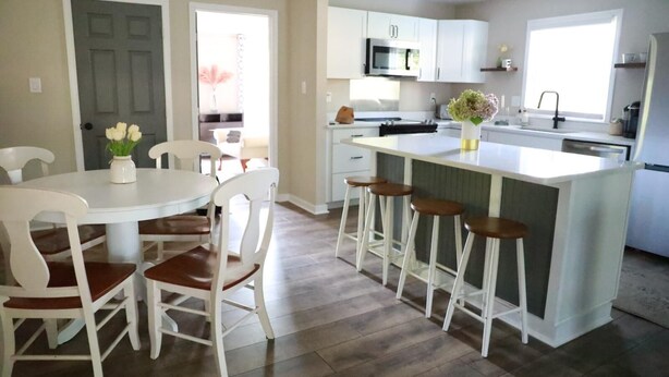 A modern kitchen with white cabinetry, a central island with stools, and a dining area with white chairs and a vase of flowers.