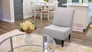 A modern living room with a glass coffee table, a gray armchair, and a dining area with white chairs and a table.