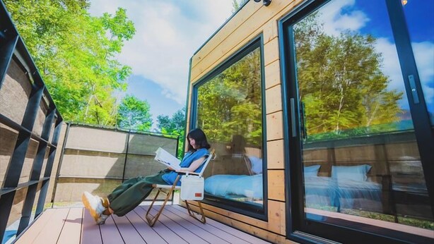 A person sitting on a deck chair reading a book in a modern room with large windows and a view of trees. - SAUNA&Villa takibi (Makkari-mura)