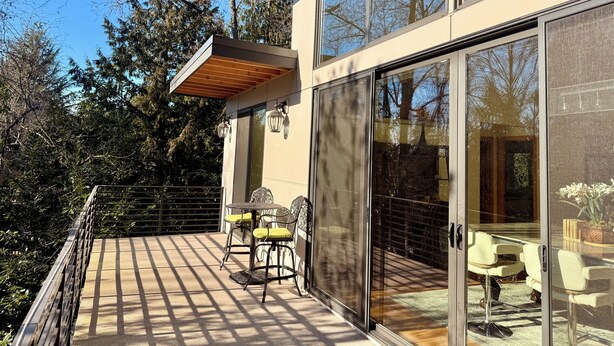 Un bâtiment moderne doté de grandes fenêtres en verre, d’une terrasse en bois avec des meubles d’extérieur et offrant une vue sur les arbres.