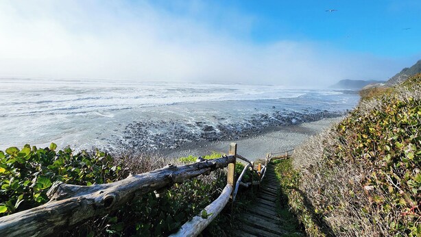 Un sendero costero con una valla de madera que conduce a una playa rocosa con olas rompiendo en la orilla.