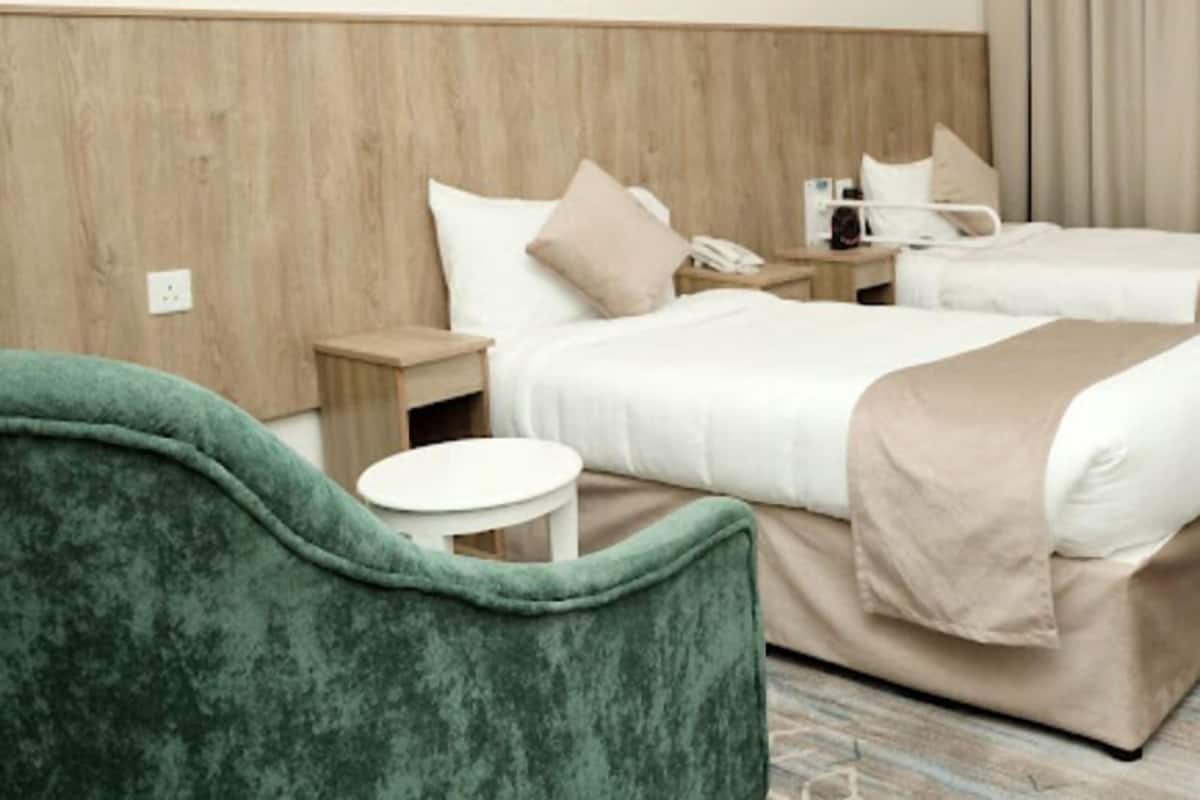 A hotel room with two beds, a green armchair, a bedside table, and a nightstand.