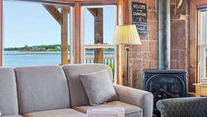 Ein gemütliches Blockhaus-Interieur mit Kamin aus Stein, einer Couch, einem gemusterten Sessel und Blick auf das Wasser.