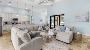 A spacious living area with a large L-shaped sofa, a round coffee table, and a dining table. There is a kitchen area with white cabinets and a microwave. The room has a ceiling fan, pendant lights, and a large window.