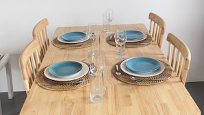 Une table à manger en bois, agrémentée d’assiettes bleues et de verres transparents, entourée de sets de table en fibres tressées et de chaises en bois.
