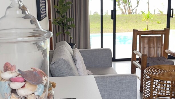 Un salon moderne avec un canapé blanc, un aquarium en verre décoré de coquillages, un fauteuil en osier et une vue sur une piscine et la verdure environnante.