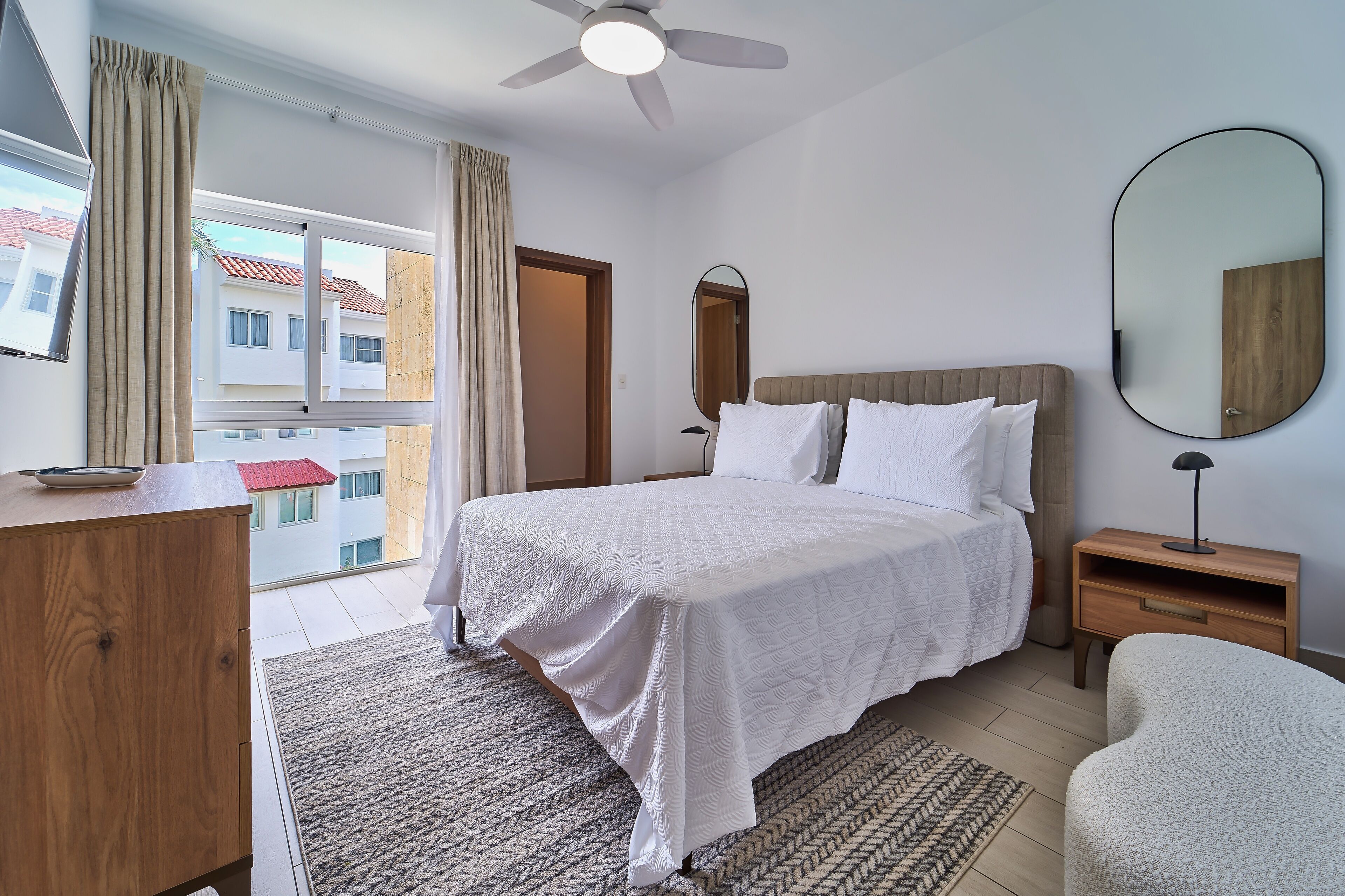 A bedroom with a bed, bedside tables, a desk, a mirror, and a view of buildings outside.