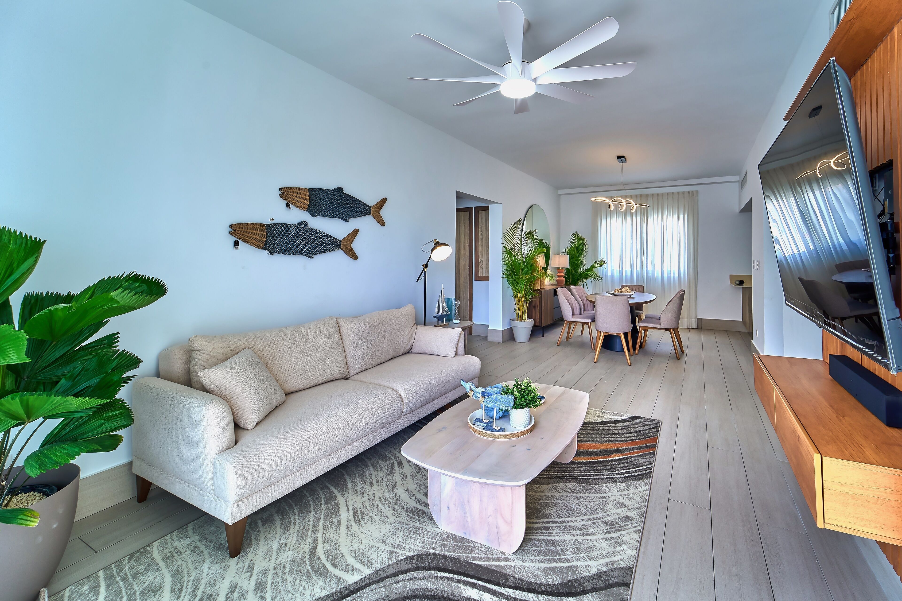 A modern living room with a beige sofa, a wooden coffee table, a ceiling fan, and a TV mounted on the wall.