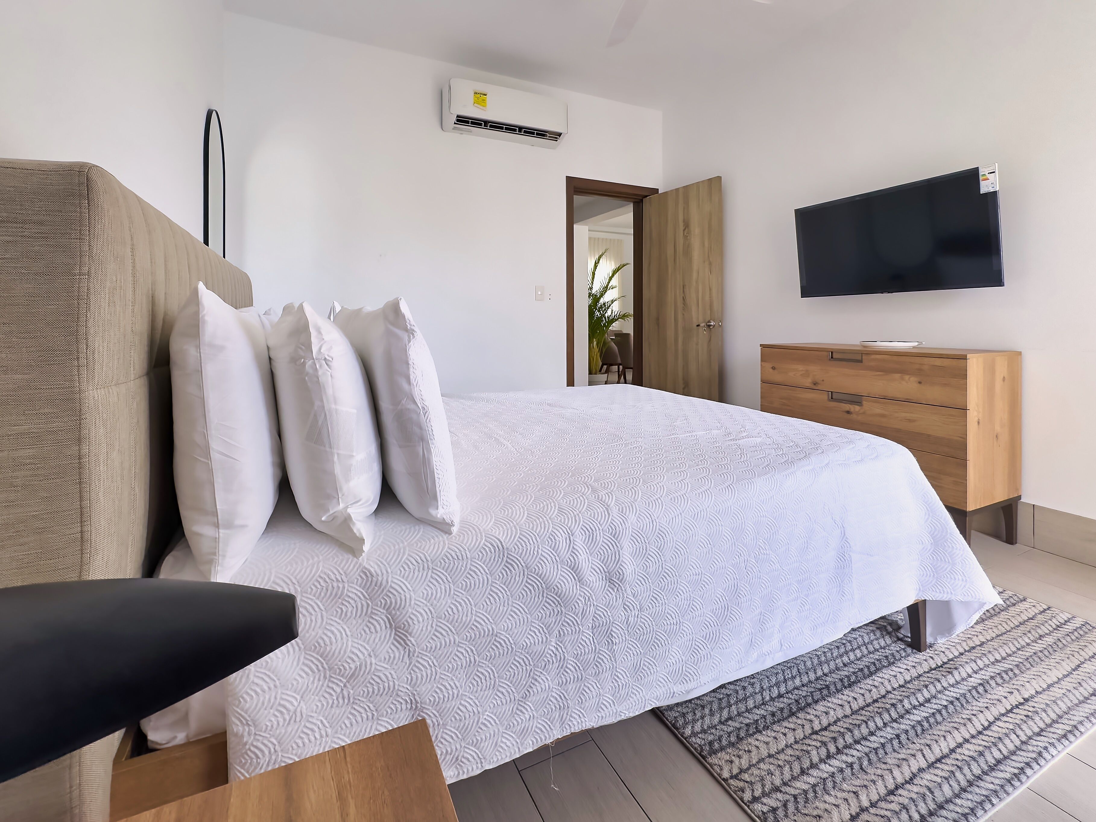 A modern bedroom with a large bed, a wooden dresser, a flat-screen TV, and a ceiling-mounted air conditioner.