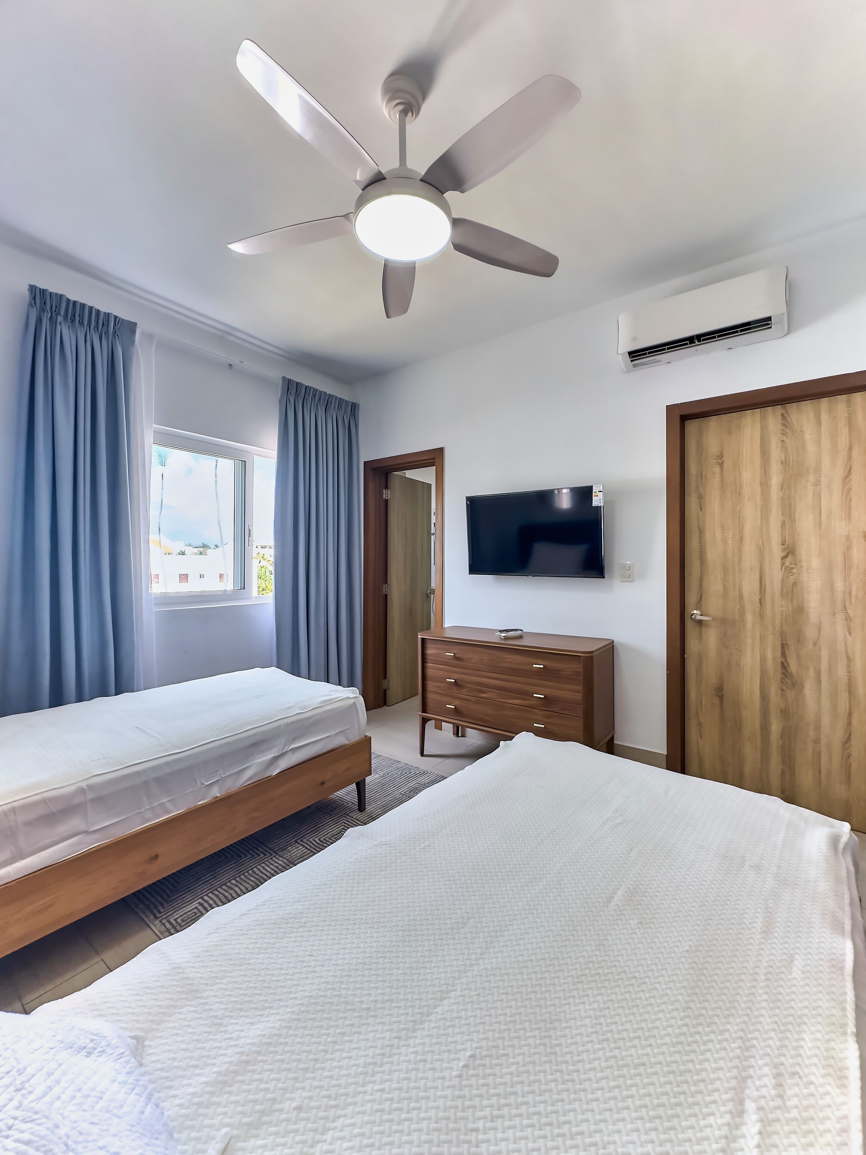 A hotel room with two beds, a ceiling fan, a TV, a wooden dresser, and a window with curtains.