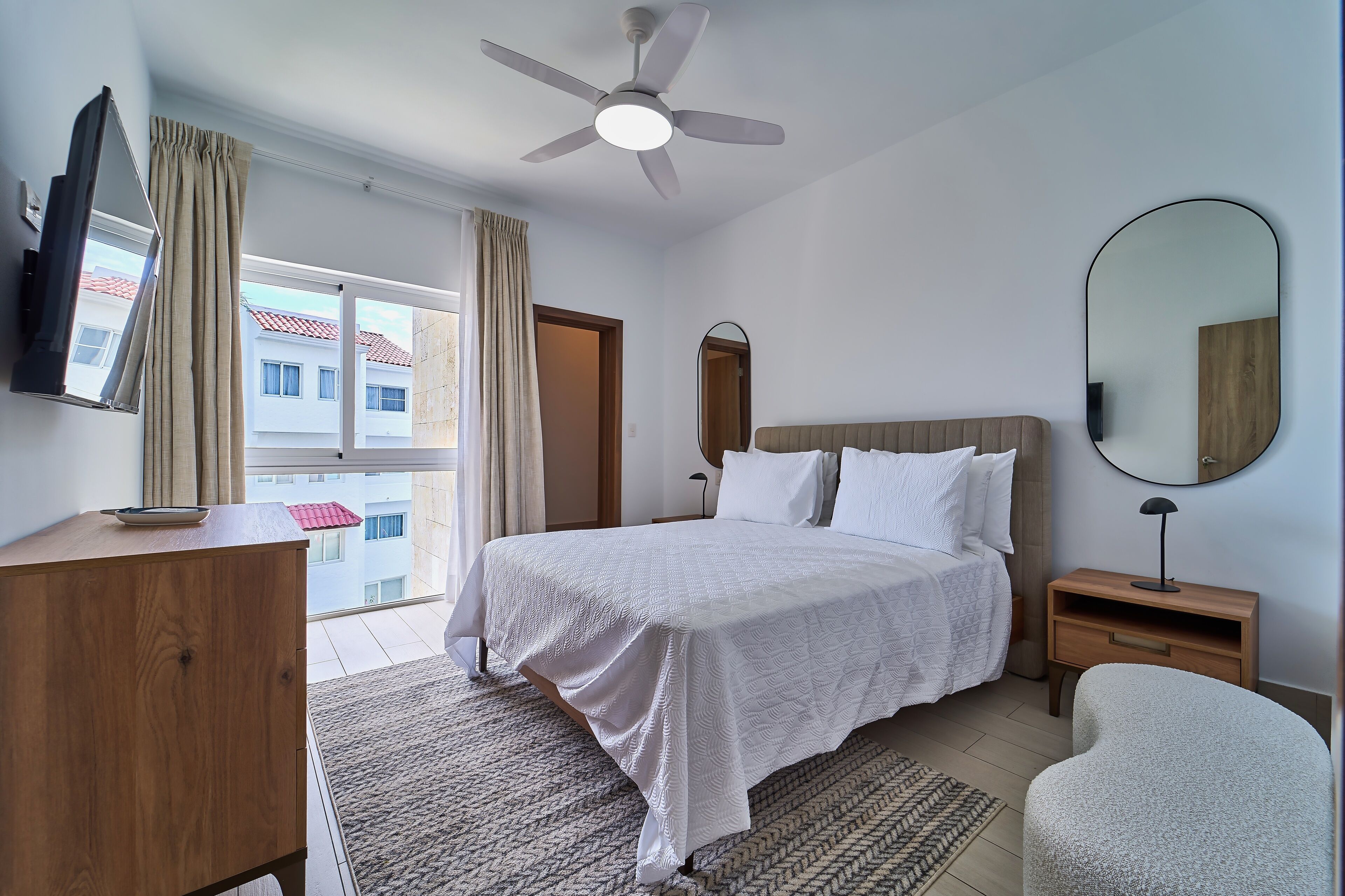 Ein Hotelzimmer mit Bett, Nachttisch aus Holz, Spiegel, Deckenventilator und Blick auf Gebäude.