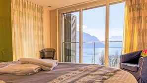 Een slaapkamer met een groot bed, een stoel en uitzicht op een meer en bergen door een schuifdeur.